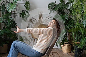 Serene relaxed young woman with closed eyes stretching arms, relaxing in urban jungle interior