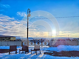 Serene Winter Sunset at Snowy Riverfront With Benches and Streetlamp. Sun sets over a tranquil, snow-covered riverbank