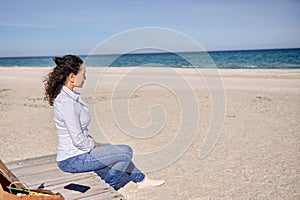 Serene woman sitting on a wooden chaise lounge sitting on lonely beach and enjoying the tranquil view to the seashore. Tranquil