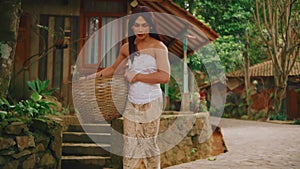 Serene woman in traditional attire holding a basket, sitting outdoors with lush greenery in the background