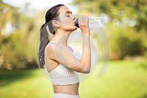 A serene young woman with her hair in a ponytail is drinking from a transparent water bottle