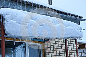 A series of icicles on the roof of the porch under the snow by the brick wall with a window