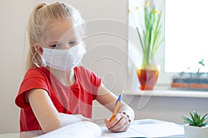 Serios sad blonde schoolgirl in medical mask stydying at home, doing school homework, writing in notebook. Reading photo