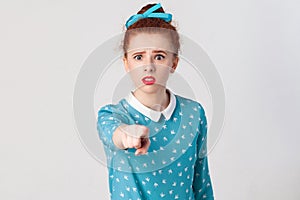 The seriosly redhead girl, wearing blue dress, opening mouths widely, having surprised shocked looks, pointing finger at camera.