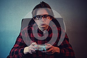 Serious adolescent boy playing video games late night seated in armchair inside a dark room background. Intent teen guy holding a photo