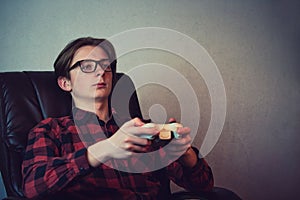 Serious adolescent boy playing video games late night seated relaxed in his armchair over grey wall background. Intent teen guy