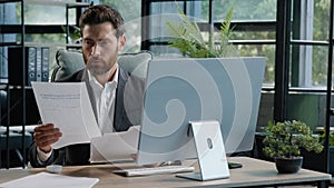 Serious adult 40s bearded pensive thoughtful businessman working at computer in office with paperwork. Professional