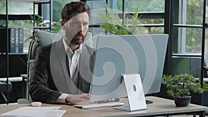 Serious adult 40s bearded pensive thoughtful businessman working at computer in office with paperwork. Professional