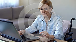 Serious adult lady in wheelchair entering card number on laptop, shopping online