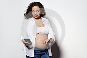 Serious adult pregnant woman with bare belly, holding smartphone and counting contractions, recording it in mobile app