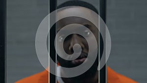 Serious African-American criminal looking at camera through prison bars, despair