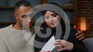 Serious African Latina man husband with arabian muslim woman wife opening envelope paper letter upset couple multiracial