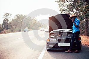 Serious african man using smartphone and holding his head by hands standing near his old broken car  with raised hood on the