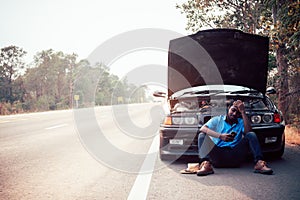 Serious african man using smartphone and holding his head by hands standing near his old broken car  with raised hood on the