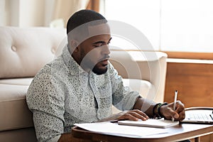 Serious african student writing notes studying at home with laptop