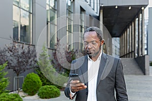 Serious afro american boss in business suit walking outside office building, mature man holding phone, businessman