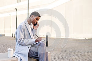 serious afro american female business woman talking by phone and taking notes on a bench