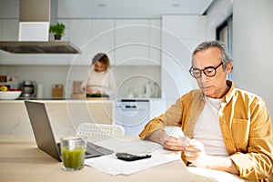 Serious aged man using cellphone, sitting at table in front of laptop in kitchen while his wife cooking on background