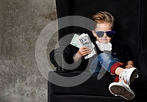 Serious arrogant rich kid boy millionaire sits with a bundle of money dollars cash in big luxury armchair