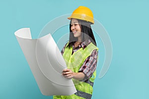 Serious asian business woman wearing yellow safety helmet holding and looking at blueprints, isolated on background in studio