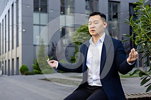 Serious Asian male businessman sitting on bench near working office in lotus position and meditating with closed eyes