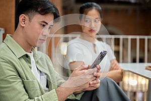 A serious Asian man is scrolling on his phone, ignoring his moody girlfriend photo