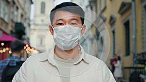 Serious asian man wearing medical mask looking at camera male tourist standing outdoors in protective respirator