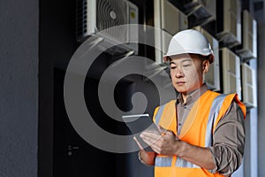 Serious Asian man works in construction and manufacturing. Standing outside in a hard hat and vest, holding a tablet