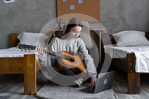 Serious asian teenager girl learning to play guitar using laptop computer sitting near the bed at home.