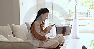 Serious Asian woman resting on modern sofa with smartphone