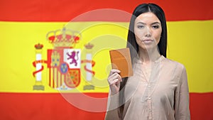 Serious Asian woman showing passport against Spanish flag background, migration