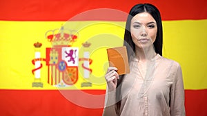 Serious Asian woman showing passport against Spanish flag background, migration