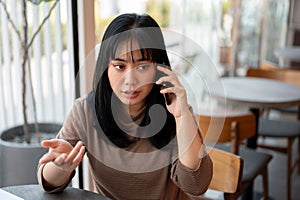 A serious Asian woman is talking on the phone with someone while sitting in a cafe