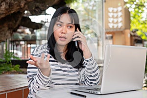 A serious Asian woman is talking on the phone with someone while working remotely at a cafe