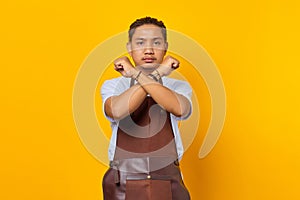 Serious asian young man showing refusal gesture with crossed arms isolated on yellow background
