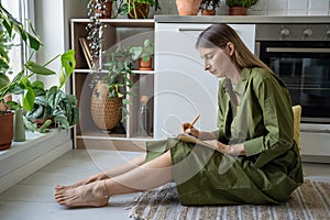 Serious attentive woman focused on notebook writing personal diary sitting on kitchen floor at home.