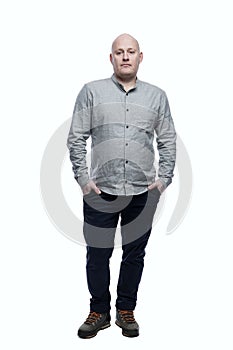 Serious bald adult man stands with his hands in his pockets. Wearing a gray shirt and blue jeans. Full height. Isolated on a white