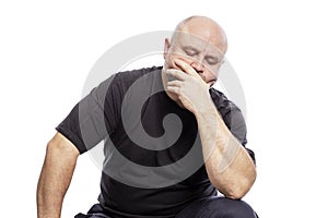 A serious bald middle-aged man in a black T-shirt is sitting with his hand in his face. Isolated over white background
