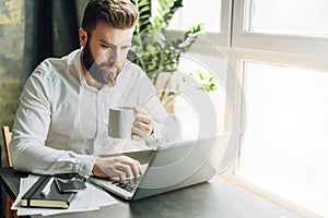 Serious bearded businessman is sitting at table, working on computer, drinking coffee. Man checking email, planning.