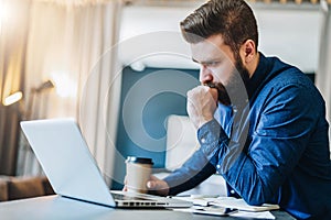 Serious bearded businessman working on computer, drinking coffee, thinking. Man analyzes information, checking email.