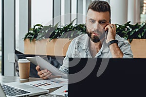 Serious bearded hipster man sits in office, talking on phone while holding tablet computer,looking at laptop screen.