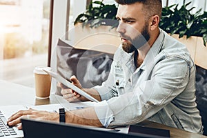 Serious bearded hipster man sitting in office at desk,working on laptop,holding tablet computer,looking at laptop screen