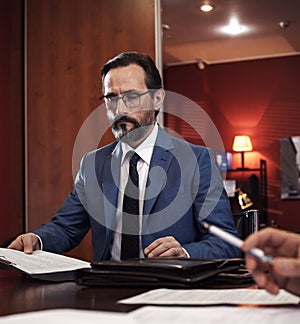 Serious bearded man in blue jacket, white shirt, tie filling documents sitting at table in office