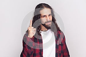 Serious bearded man in checkered red shirt raises finger up, warning, has confident look.