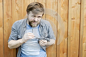 Serious bearded man cleans glasses by rubbing them with the help of his shirt and squinting, looking at the camera with hostility