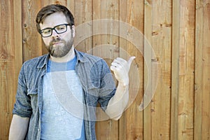 A serious, bearded man with glasses shows his thumb on a wooden, old wall and looks away. Isolated