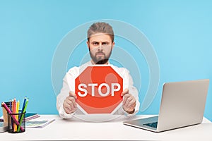 Serious bearded man office worker holding red stop sign board sitting at workplace with laptop, rejecting sexual harassment at