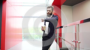 Serious bearded man is standing with cup of take-out coffee near glass fence, looking at watch and waiting for someone