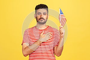 Serious bearded man in striped t-shirt holding united states of america flag holding hand on heart, swearing allegiance,