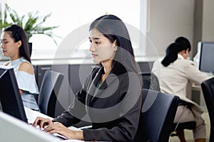 Serious beautiful Asian woman officer typing desktop computer while sitting at office desk with blurred background of her busy